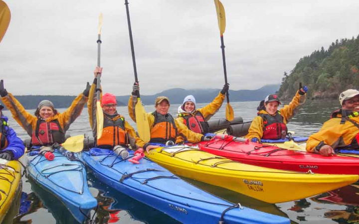a group of kayakers raise smile and their paddles in the air on a sea kayaking trip with outward bound
